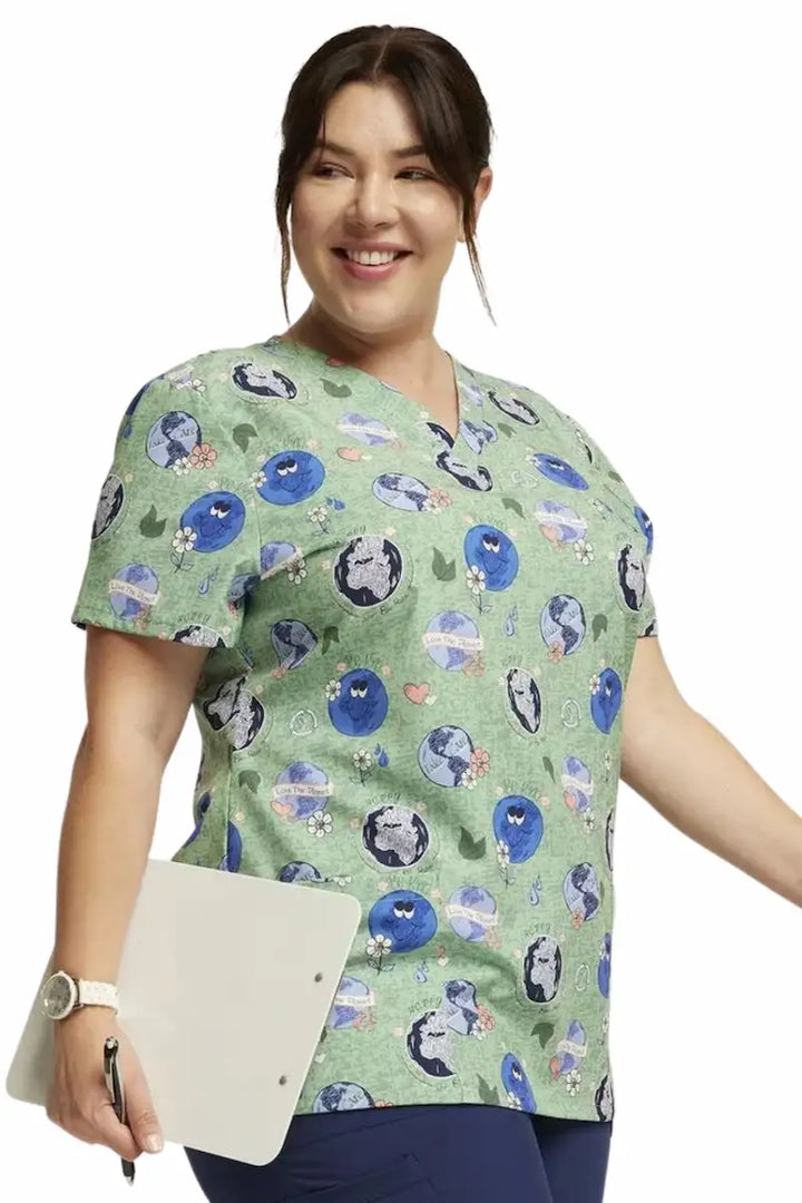 A nurse displaying the right side of the Dickies Women's V-Neck Print Scrub Top in Happy To Be Here featuring short sleeves and a v-neckline.