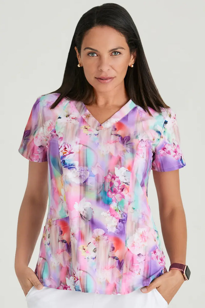 A young female Pediatric Nurse showcasing the front of the Barco One Women's Print V-Neck Scrub Top in Floral Blooms featuring a soft, 4-way stretch fabric that is breathable and durable.