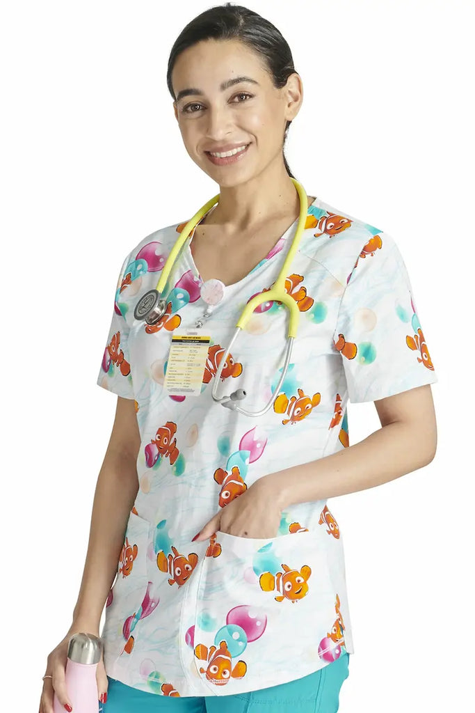 A young female Pediatric Nurse wearing a Tooniforms Women's V-neck Printed Scrub Top in "Nemo Bubbles" in size 2XL featuring a Modern Classic Fit.
