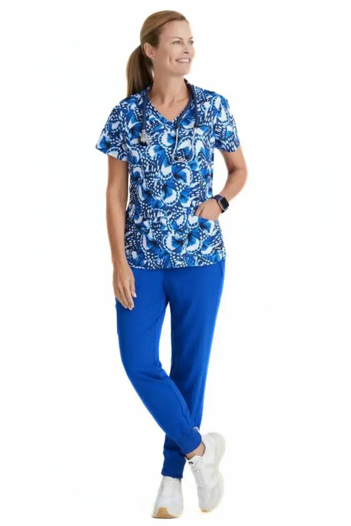 A young female Psychiatric Nurse wearing a Grey's Anatomy Women's Printed Scrub Top in "Butterfly Blues" featuring a vibrant butterfly print on a soft, stretchy spandex fabric that moves with you throughout your busy shift.