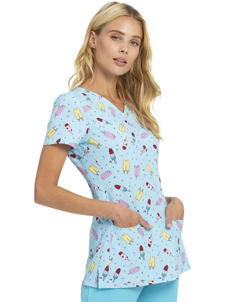 A young female Pediatrician wearing a HeartSoul Women's V-neck Print Scrub op in "Popsicle Party" size Large featuring front & back princess seams for shaping.