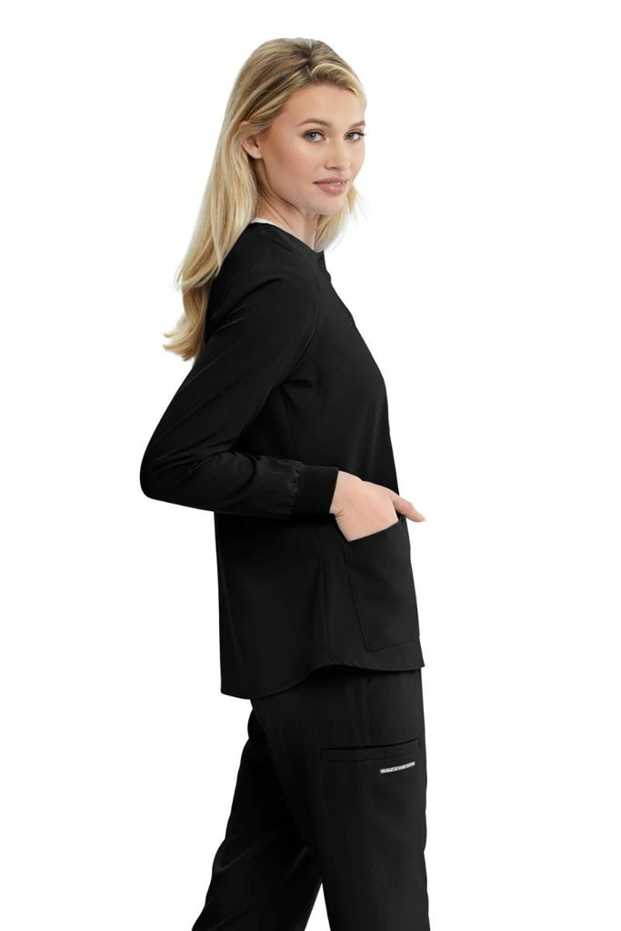 A young female physical therapist showcasing the right side of the Skechers Women's Stability Scrub Jacket in Black featuring slanted shoulder yoke seaming.