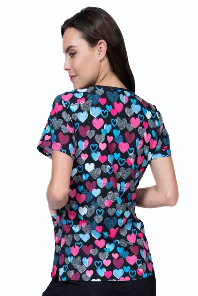 A young lady nurse wearing a Meraki Sport Women's Print Scrub Top in "Follow Your Heart" featuring shoulder yokes & side slits for additional range of motion.