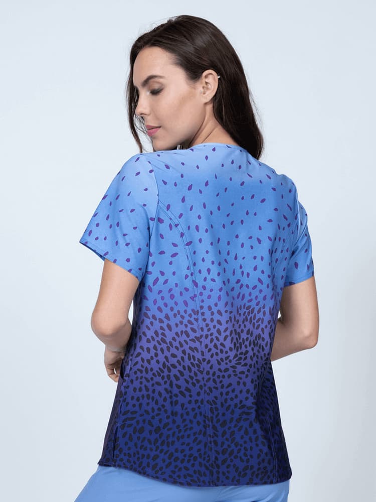 A young female Nurse Practitioner wearing a Women's Print Scrub Top from Meraki Sport in "Grape Sensation" featuring shoulder yokes & side slits for additional range of motion.