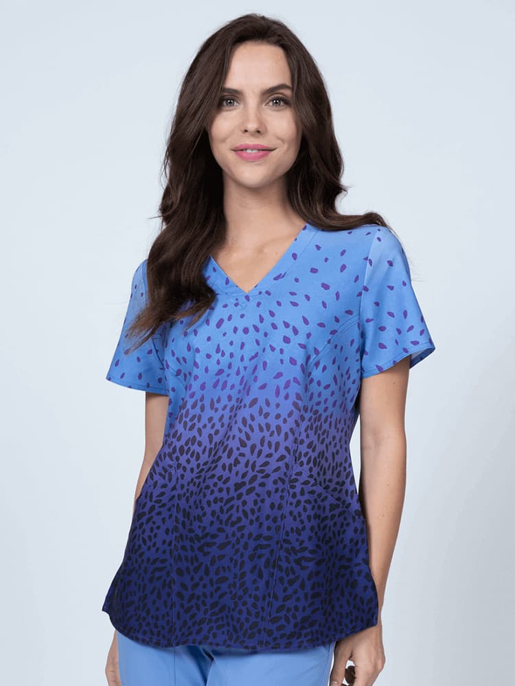 A young female nurse wearing a Meraki Sport Women's Print Scrub Top in "Grape Sensation" featuring a v-neckline & short sleeves.