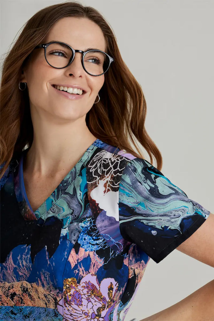 A young female children's nurse wearing a Barco one Women's Printed Scrub Top in Mystic Flower featuring short sleeves and stylish seaming throughout.