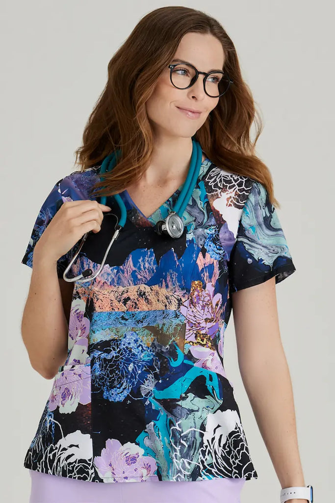 A young female Pediatric Nurse wearing a Barco One Women's Print V-neck Scrub Top in Mystic Flower featuring two spacious front pockets.