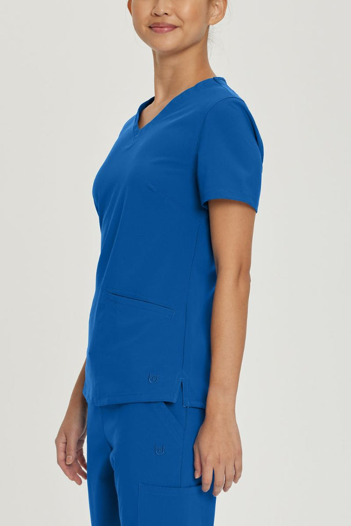 A young female Nursing Assistant wearing an Urbane Performance Women's Motivate V-neck Scrub Top in Royal Blue size XL featuring side slits with mesh detail to provide additional range of motion and added comfort.