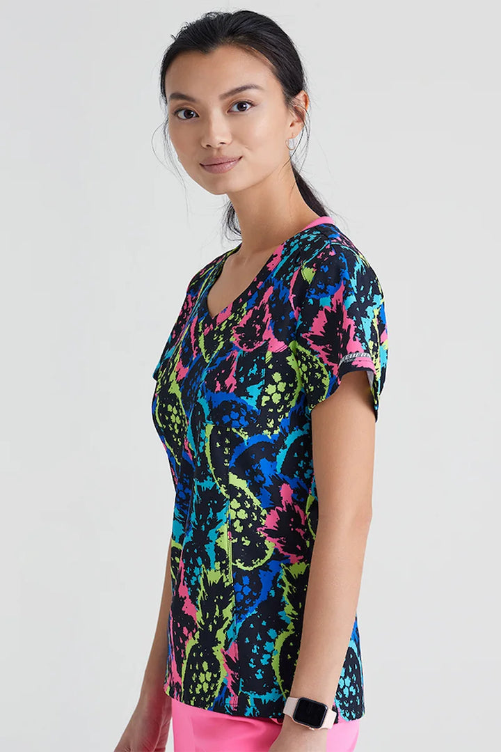 A young female Pschiatric Nurse wearing a Skechers Women's Printed Scrub Top in Pineapple Pop featuring side venst for enhanced breathability throughout the day.