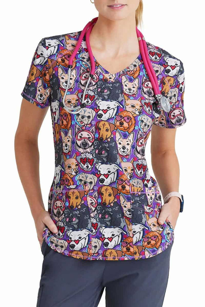 A young female Veterinarian wearing a Skechers BOBS Women's Printed Scrub Top in "Part of the Family" featuring various breeds of dogs wearing sunglasses on a purple background.