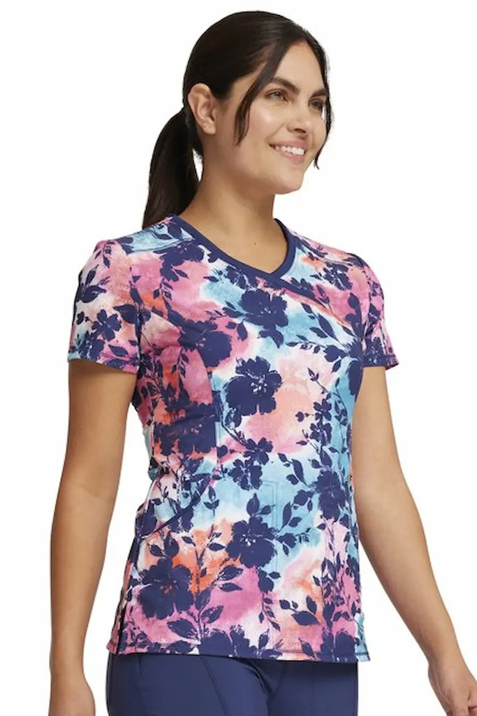 A young female LPN wearing a Cherokee Infinity Women's Mock Wrap Printed Scrub Top in "Artistic Blooms" size XS featuring intricate floral patterns and vivid colors, adding a touch of personality and elegance. 
