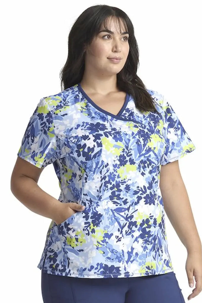 A young female Pediatric Nurse wearing a Cherokee Infinity Mock Wrap Printed Scrub Top in "Brushstroke Buds" size XL featuring 2 set-in pockets.