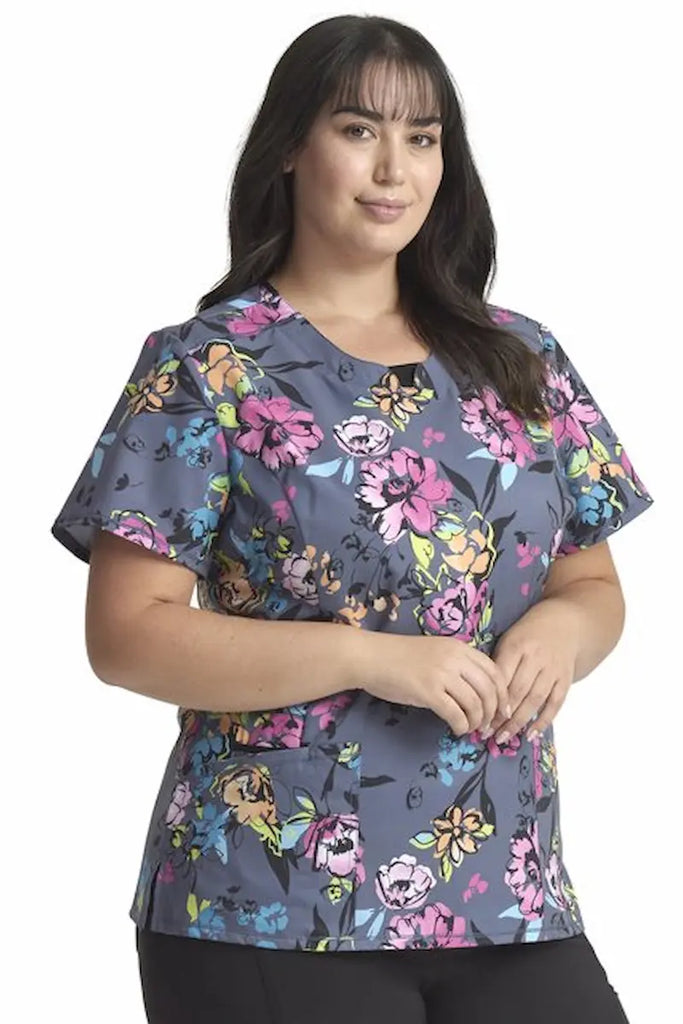 A young female Labor and Delivery Nurse wearing a Cherokee Infinity Women's Round Neck Printed Scrub Top in "Electric Blossoms" size Small featuring two front patch  pockets.
