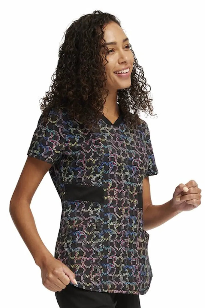 A young female LPN wearing a Cherokee Infinity Women's V-Neck Printed Scrub Top in "Links of Love" featuring side slits for additional range of motion throughout the day.