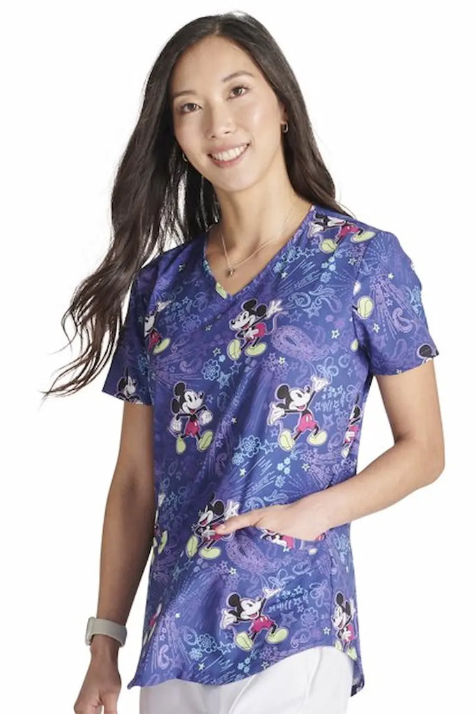 A young female Childcare Specialist wearing a Tooniforms Women's V-neck Printed Scrub Top in Mickey's Bandana Land size Medium featuring a soft and breathable fabric made of 55% cotton and 45% polyester.