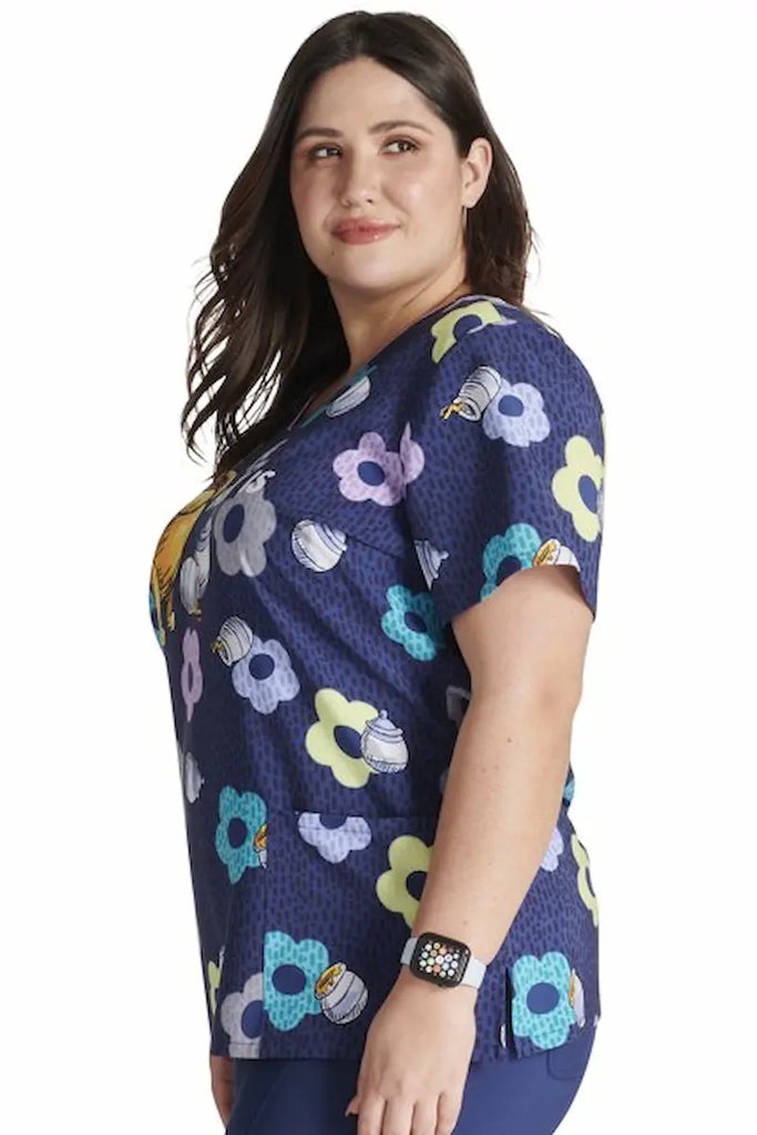 A young female Psychiatric Nurse wearing a Tooniforms Women's V-neck Printed Scrub Top in "Home & Hunny" featuring an adorable design with Winnie the Pooh surrounded by honey bees and flowers, adding a touch of sweetness and charm.