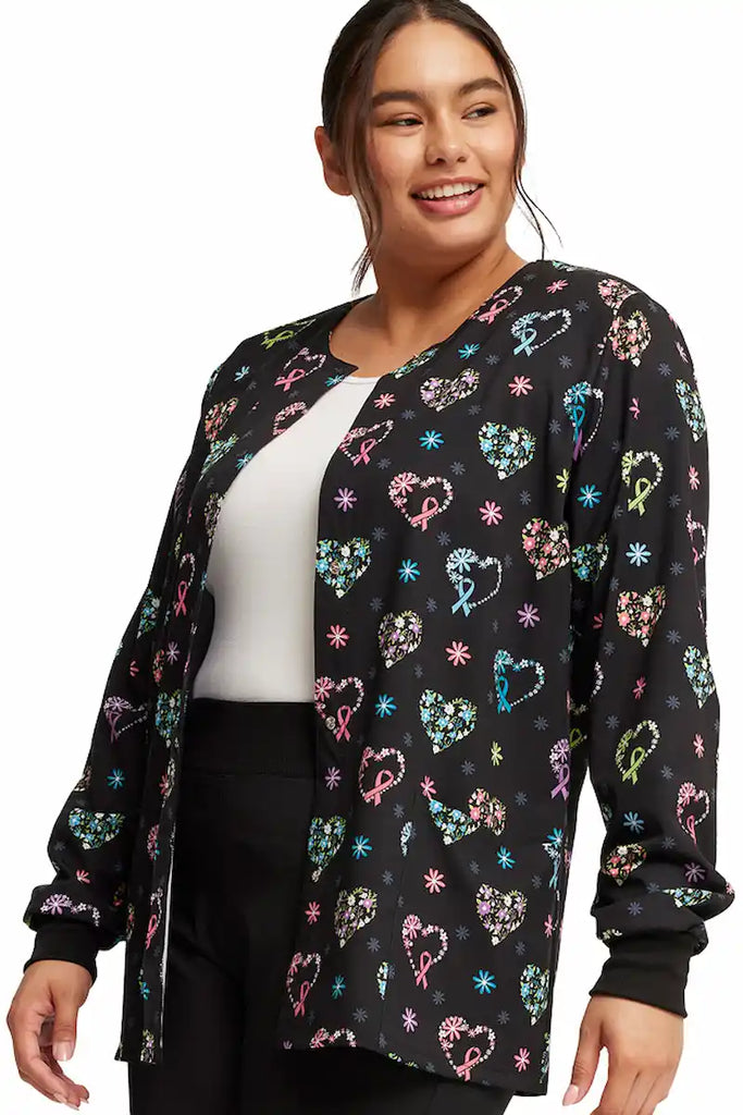 A young female Neonatal Nurse wearing a Cherokee Women's Printed Scrub Jacket in "Care Flor-All" featuring a round neckline and soft & breathable fabric made of 92% Polyester and 8% Spandex.