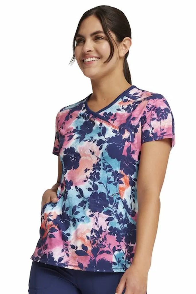 A young female Oncology Nurse wearing a Cherokee Infinity Women's Mock Wrap Scrub Top in "Artistic Blooms" size small featuring 2 on-seam pockets for ample storage space.