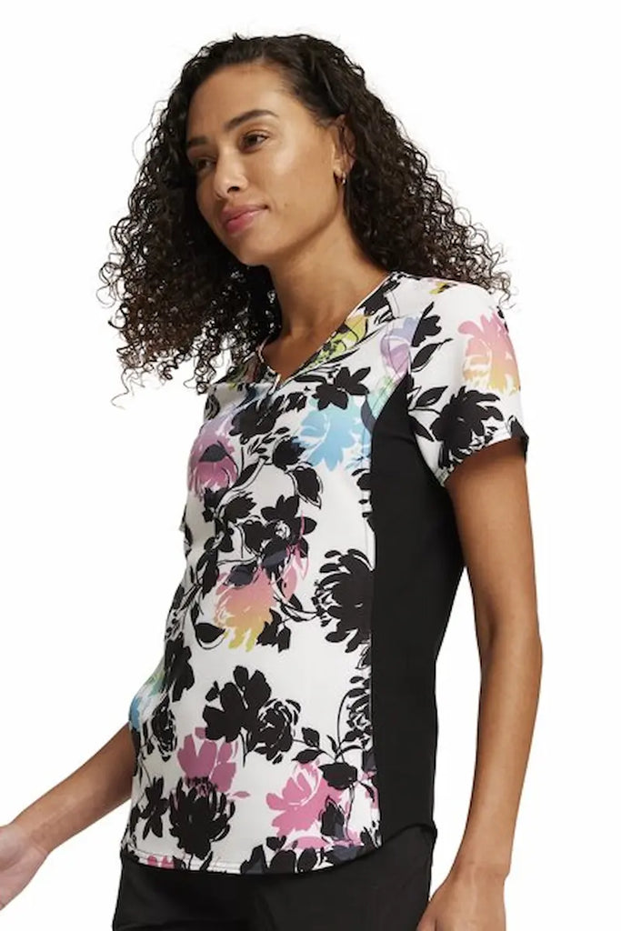 A young female Chilcare SPecialist wearing a Cherokee iFlex Women's V-neck Printed Scrub Top in "Garden Glow" size 2XL featuring knit side panels for a comfortable all day fit.