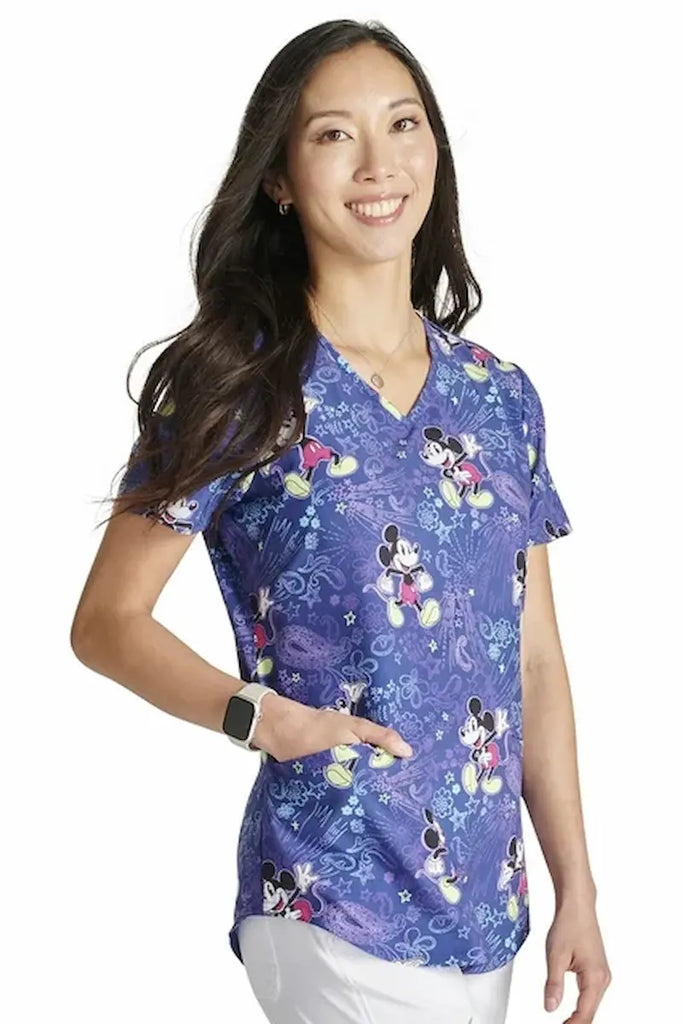 A young female Pediatric Nurse Wearing a Tooniforms Women's V-neck Printed Scrub Top in "Mickey's Bandana Land featuring two front patch pockets for ample storage space.