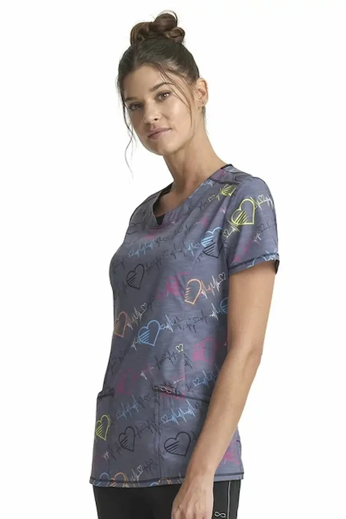 A young female Neonatal Nurse wearing a Cherokee Infinity Women's Round Neck Printed Scrub Top in "Pop Beats" size Large featuring two front angled patch pockets.