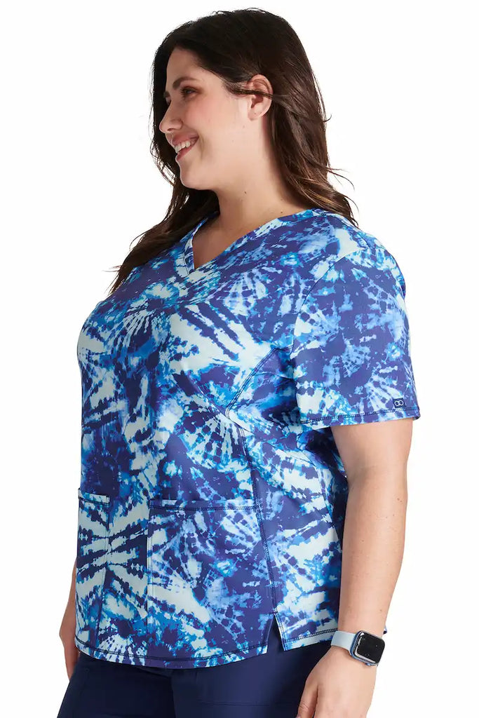 A young female Psychiatric Nurse wearing a Cherokee Women's V-neck Print Scrub Top in "Tie Dye Tranquility" size XS featuring side vents for additional range of motion.