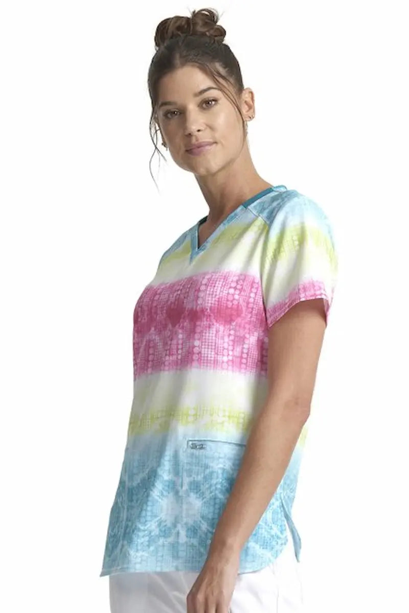 A young female Pediatric Nurse wearing a Cherokee Women's V-neck Printed Scrub Top in "Trippy Stripes" featuring a shirttail hemline & side slits for a flattering and comfortable all day fit.