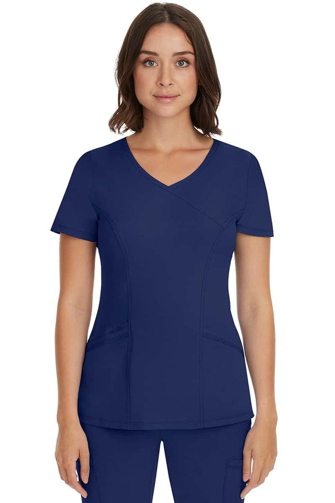 A young female Registered Nurse wearing an HH-Works Women's Madison Mock Wrap Scrub Top in Navy featuring a faux wrap neckline.