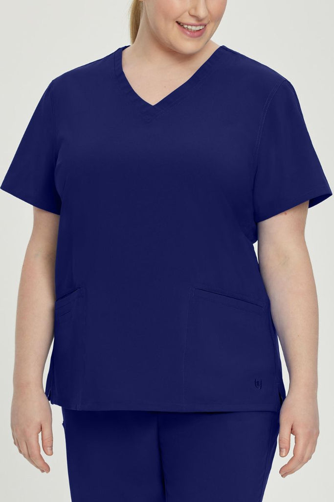 A young female CNA wearing an Urbane Performance Women's Motivate V-neck Scrub Top in True Navy size 2XL featuring tonal stitching and bust darts.