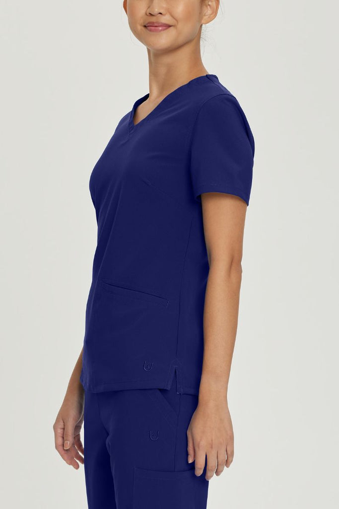 A young female nurse wearing an Urbane Performance Women's Motivate V-neck Scrub Top in True Navy size large featuring side slits with mesh detail to provide additional range of motion and added comfort.