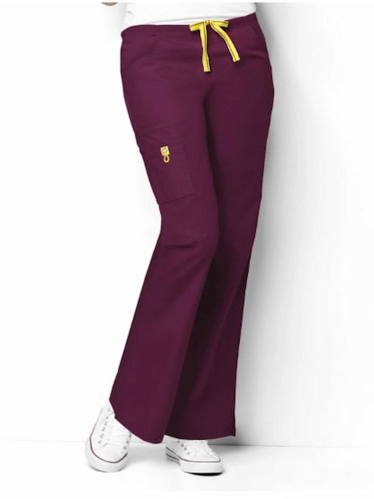 A middle aged female Nursing Assistant wearing a WonderWink Origins Women's Romeo Flare Leg Scrub Pant in Wine size Medium featuring a flat front with convertible signature yellow twill tape drawstring and back elastic waistband.