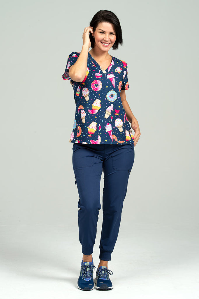 A young female Home Health Care Aide wearing a Meraki Sport Women's Print Scrub Top in "Sweets & Treats" size Small featuring a unique 4-way stretch fabric designed to move with you all day.