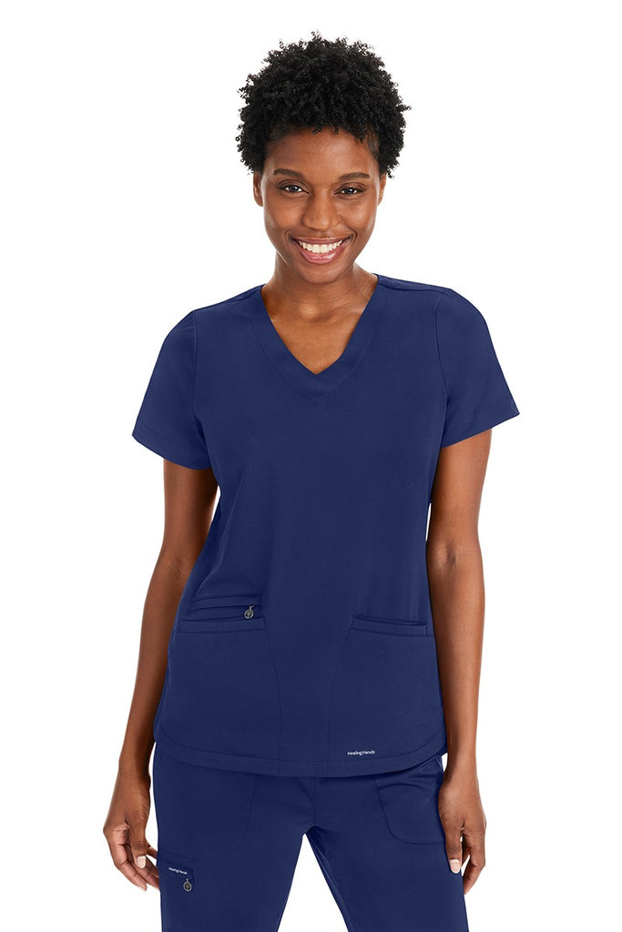 A young female Nurse Practitioner wearing a Purple Label Women's Andes Curved V-Neck Scrub Top in "Navy" size XS featuring 2 front patch pockets.