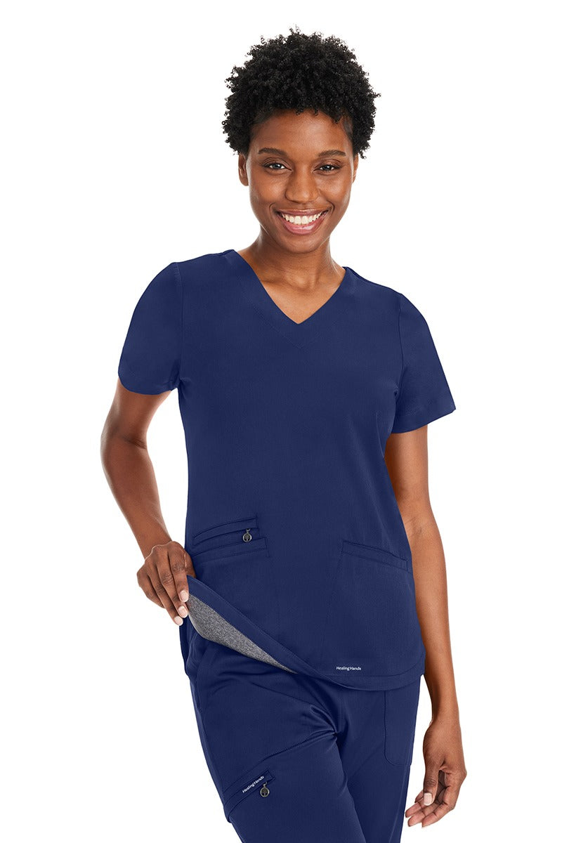 A young female Pharmacy Technician wearing an Andes Curved V-Neck Scrub Top from Purple Label by Healing Hands in "Navy" size Medium featuring an added layer of knit fabric throughout.