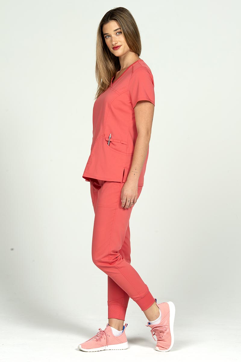 A full body image of a female Nurse Practitioner wearing an Epic by MedWorks Women's Y-Neck Scrub Top in Coral size large featuring a total of 4 pockets.