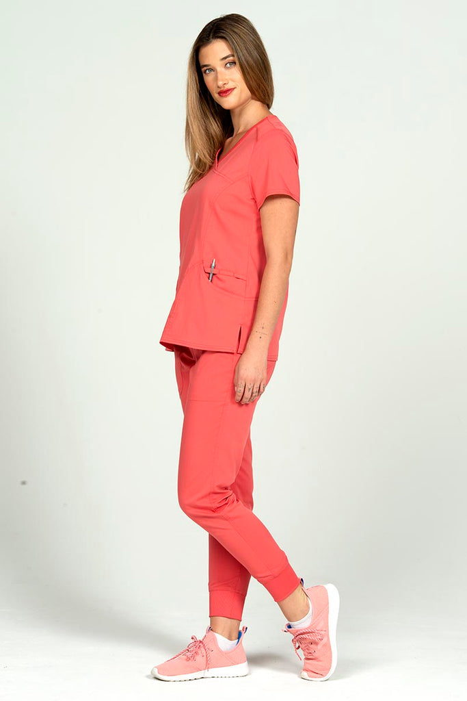 A full body image of a female Nurse Practitioner wearing an Epic by MedWorks Women's Y-Neck Scrub Top in Coral size large featuring a total of 4 pockets.