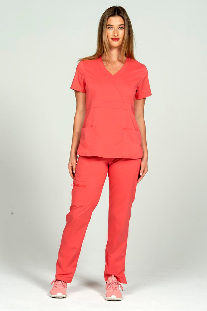 A full body image of a young nurse wearing a Women's Mock Wrap Scrub Top from Epic by MedWorks in Coral size XS featuring a Y-neckline & short sleeves.