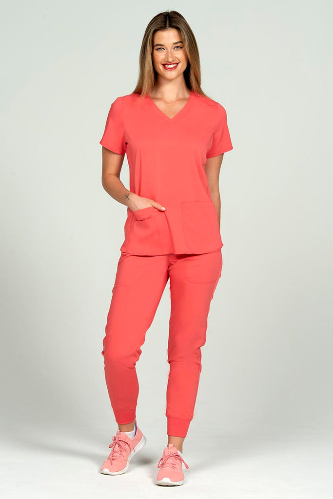 A young female home health aide wearing an Epic by MedWorks Women's Blessed Scrub Top in Coral size medium featuring stylish seaming throughout to ensure a flattering fit.
