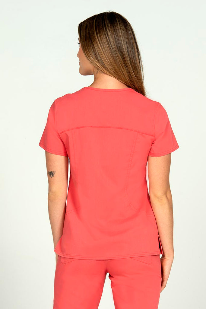 A young Home Health Aide wearing an Epic by MedWorks Women's Knit Collar Mock Wrap Scrub Top in Coral size medium featuring stylish seaming throughout.