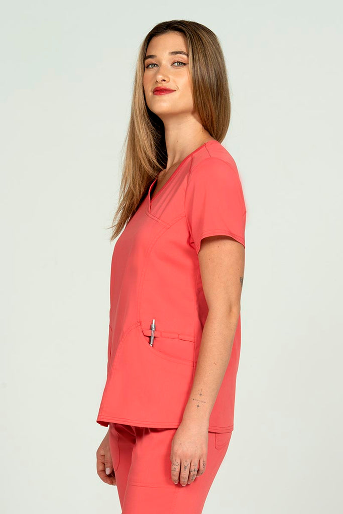 A young woman wearing an Epic by MedWorks Women's Knit Collar Mock Wrap Scrub Top in Coral featuring an easy care, 2 way stretch fabric that allows air to pass easily through the garment.