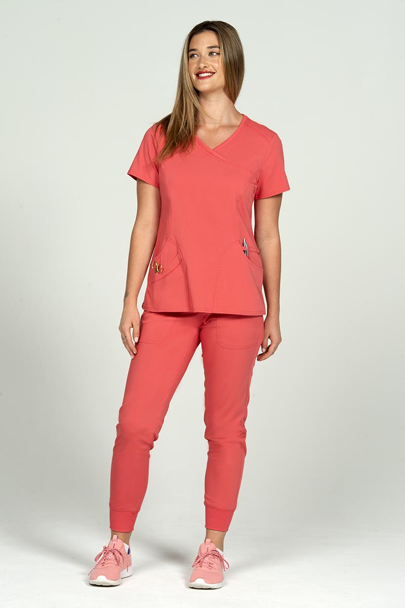 A full body image of a young female Nurse Practitioner wearing an Epic by MedWorks Women's Knit Collar Mock Wrap Scrub Top in Coral size XL featuring a total of 4 pockets.