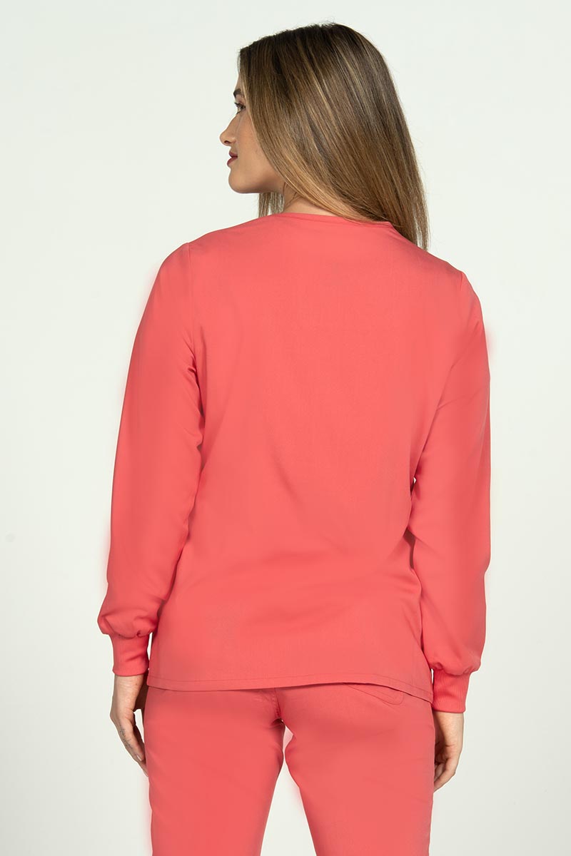 A young female Nurse Practitioner wearing an Epic by MedWorks Women's Snap Front Scrub Jacket in Coral size small featuring a tapered cut for a tailored look without losing mobility.