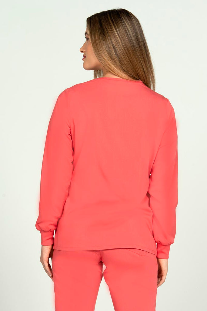 A young female Nurse Practitioner wearing an Epic by MedWorks Women's Snap Front Scrub Jacket in Coral size small featuring a tapered cut for a tailored look without losing mobility.