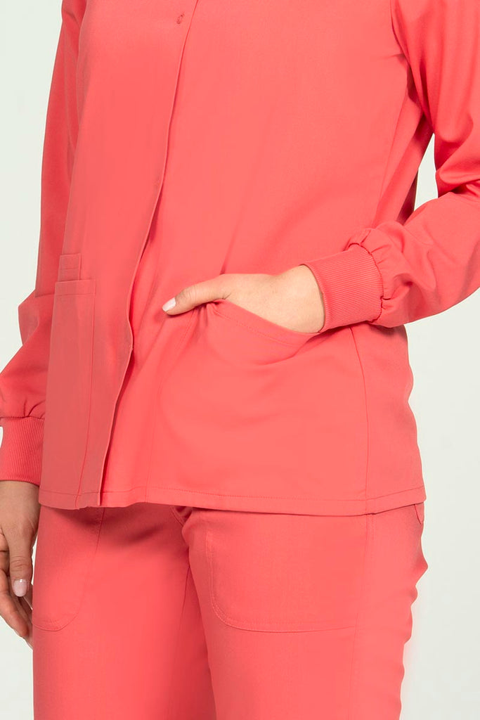A young Home Care Registered Nurse wearing an Epic by MedWorks Women's Snap Front Scrub Jacket in Coral size XS featuring 2 front patch pockets.