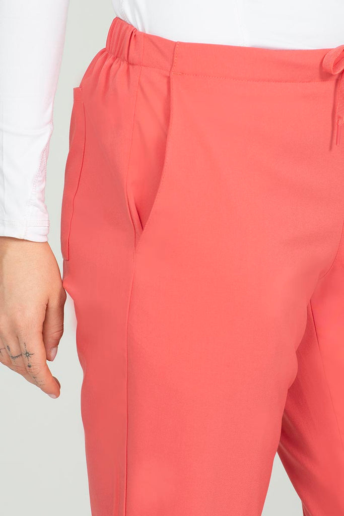 A young female Dental Assistant wearing an Epic by MedWorks Women's Natural Rise Flare Leg Scrub Pant in Coral size XS Petite featuring two front slash pockets.