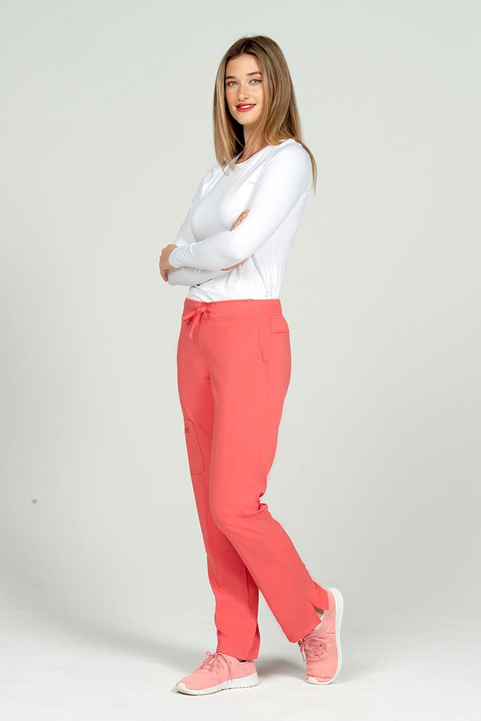 A full body image of a young female Cardiovascular Technologist wearing an Epic by MedWorks Women's Skinny Yoga Scrub Pant in Coral size XL featuring a knit waistband with an adjustable drawstring.