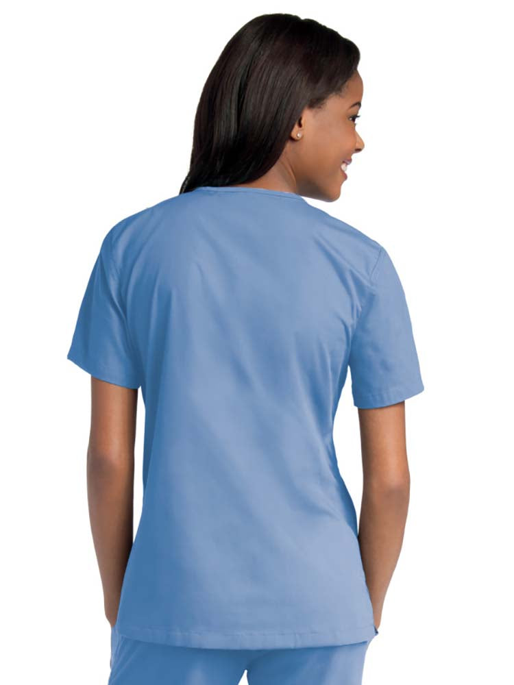 Young female healthcare worker wearing an Urbane Essentials Women's Crossover Scrub Top in "Ceil" featuring side vents for increased range of motion.
