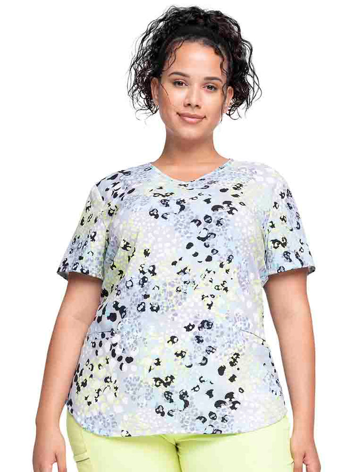 Young female healthcare worker wearing a Cherokee Women's V-Neck Print Scrub Top in "Spots Go" featuring a curved hemline.