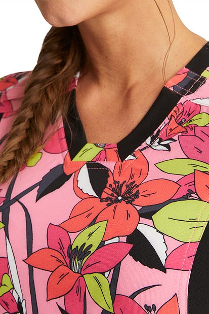 A young female nurse wearing a Cherokee iFlex V-Neck Knit Panel Print Scrub Top in "Retro Blooms" featuring a V-neckline with contrast stretch knit insets.