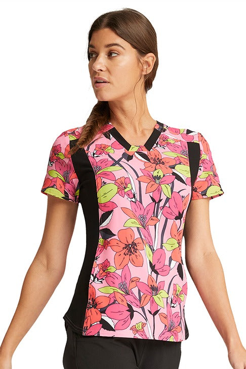 A young female LPN wearing a Cherokee iFlex V-Neck Knit Panel Print Scrub Top in "Retro Blooms" featuring a contrast stretch knit side panels.